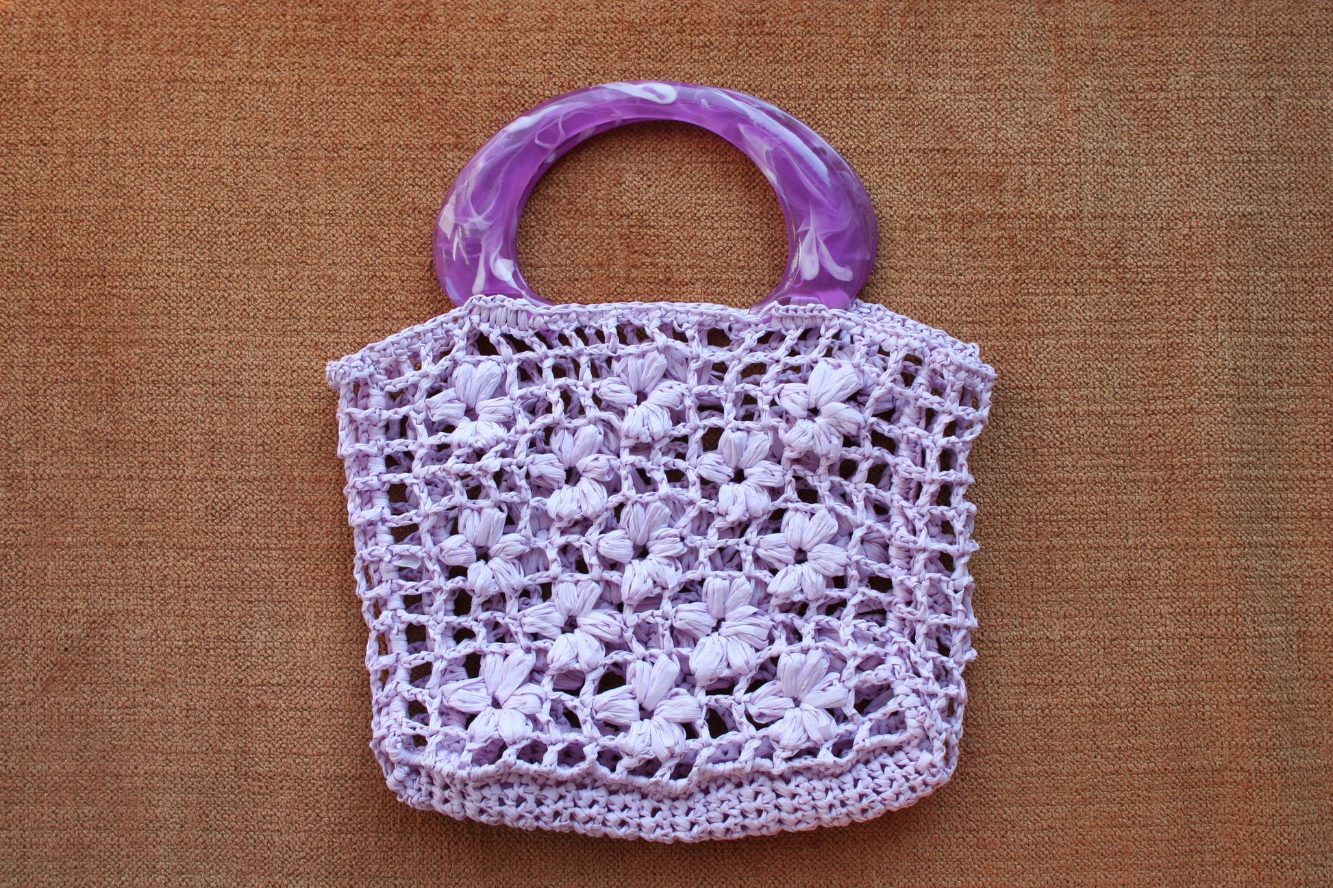 The jazmin raffia bag in lilac laid flat on a couch