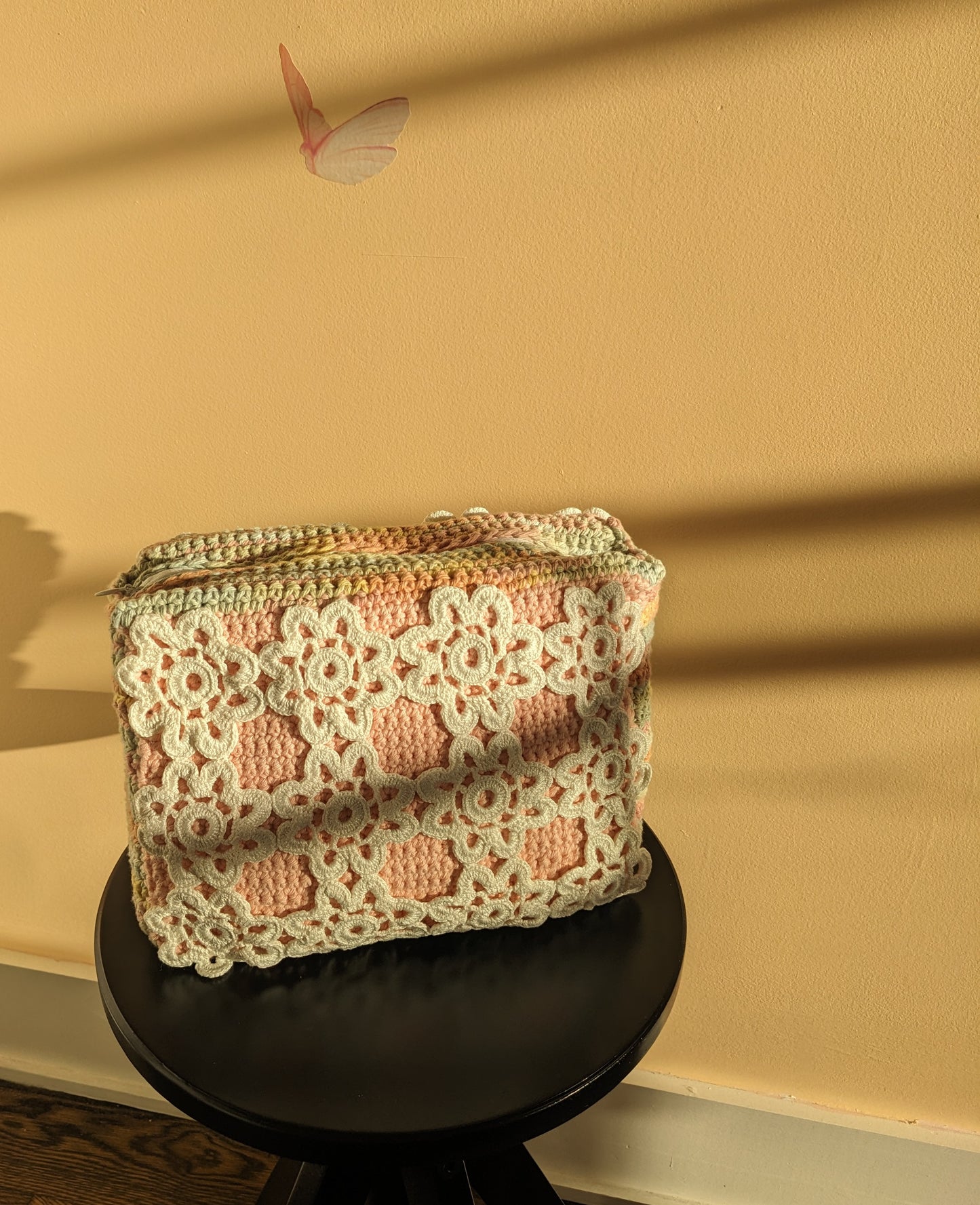 Crocheted pouch with floral pattern on a black stool against a salmon wall.